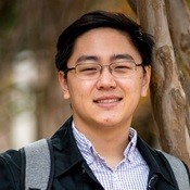 Ben Chi is a Chinese American man with short black hair. He is wearing glasses, a white and grey striped button-up, and a dark blue jacket. A shoulder strap for a satchel or backpack can be seen on his right shoulder.