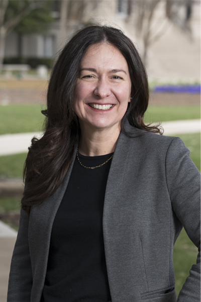 A woman stands outdoors on a college campus, smiling confidently at the camera. She has long, dark brown hair worn loose, and is dressed in a charcoal gray blazer over a black top, accessorized with a simple gold necklace. The background shows a grassy lawn, pathways, and academic buildings.