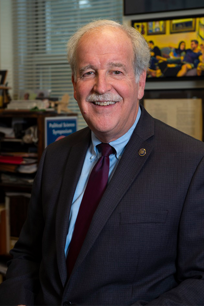 Dr. Kobylka is a white man with short, light-colored hair wearing a dark suit jacket, light blue dress shirt, and burgundy tie. He is smiling while seated in an office with books, papers, and framed artwork visible in the background.