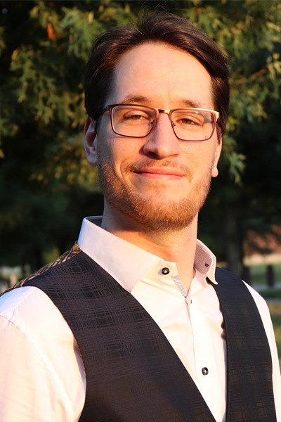 Dr. Nils Van den Steen is a white man with glasses, short brown hair, and a short beard and mustache. He is dressed in a white button-down shirt and dark plaid patterned vest, standing outside in front of trees