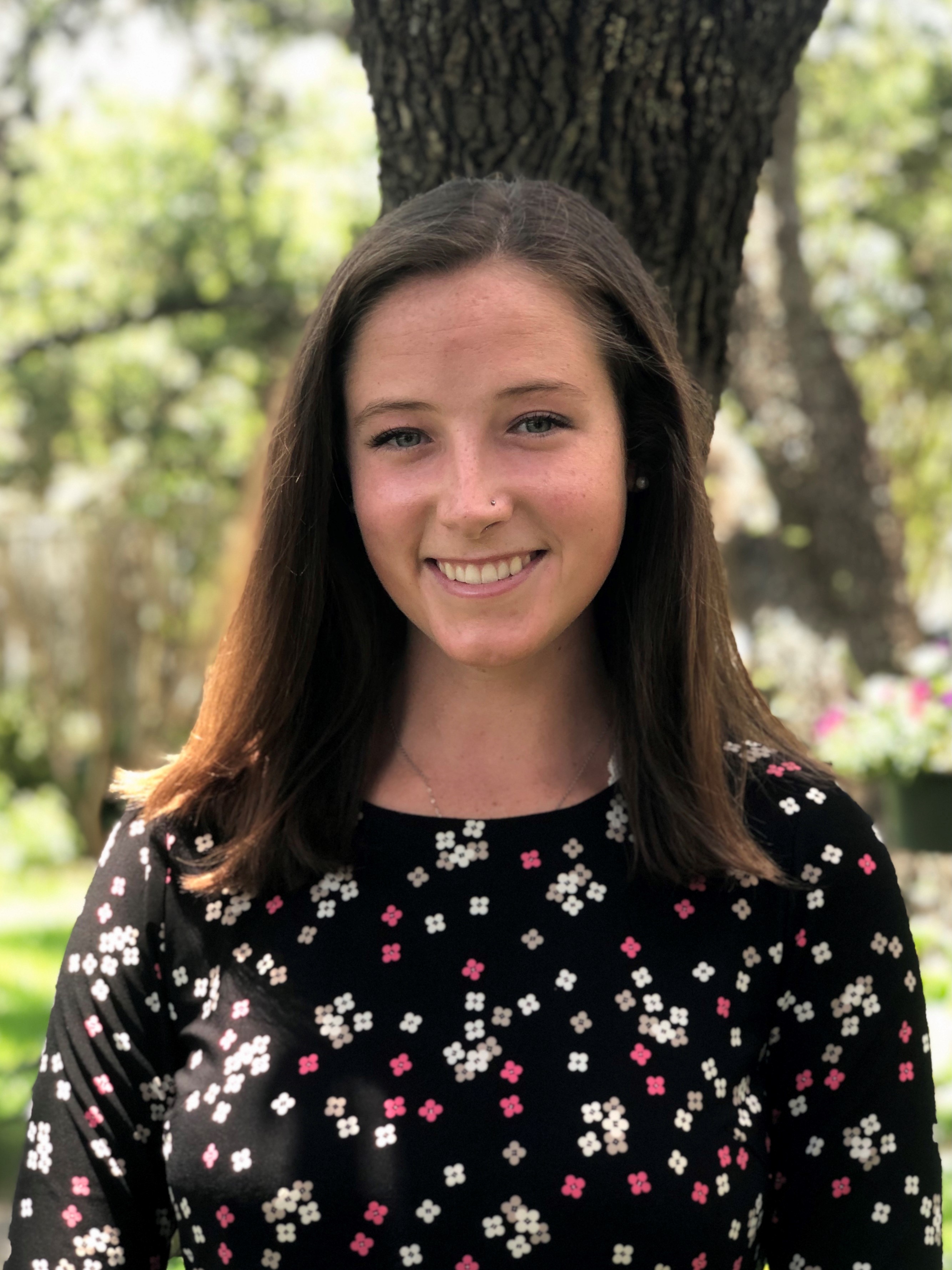 Isabelle Galko is a white woman with light brown hair approximately to her shoulders. She is wearing a black dress with white and pink flowers, and is standing in front of a tall tree.