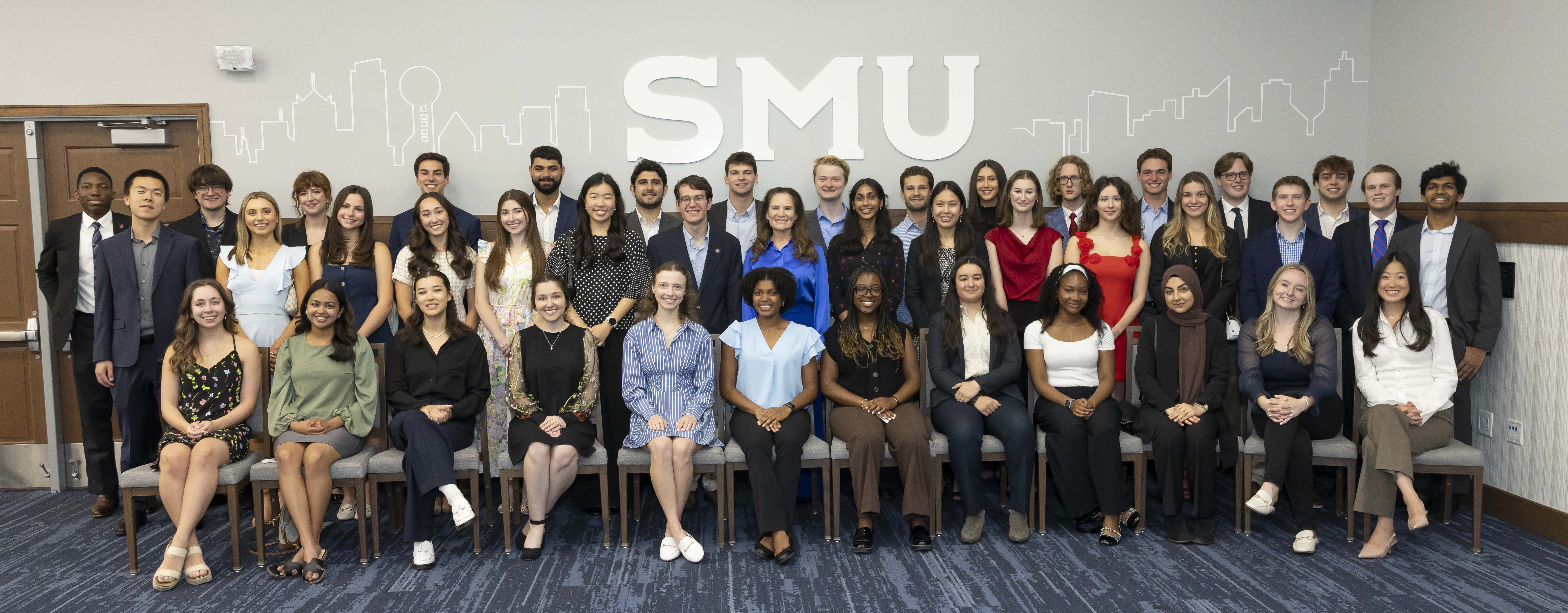 Approximately 40 students arranged in 2 rows of standing students and 1 row of sitting students pose in front of the SMU logo with the Dallas skyline decal