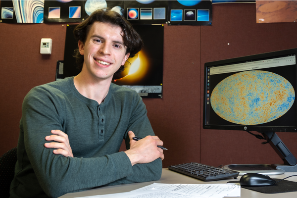 SMU's first Churchill Scholar, Joshua Ange,  sits at a desk with arms crossed in front of a computer monitor displaying a colorful cosmic map, with astronomy images pinned on the wall behind them and papers spread across the desk.