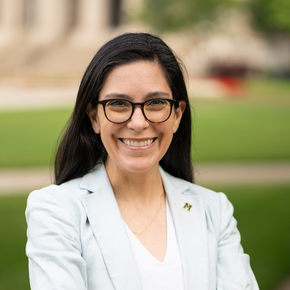 Caitlin is a white woman with dark brown hair. She is wearing black framed glasses, a light blue blazer, and a white v-neck blouse underneath. The background is the blurred path to Dallas Hall.