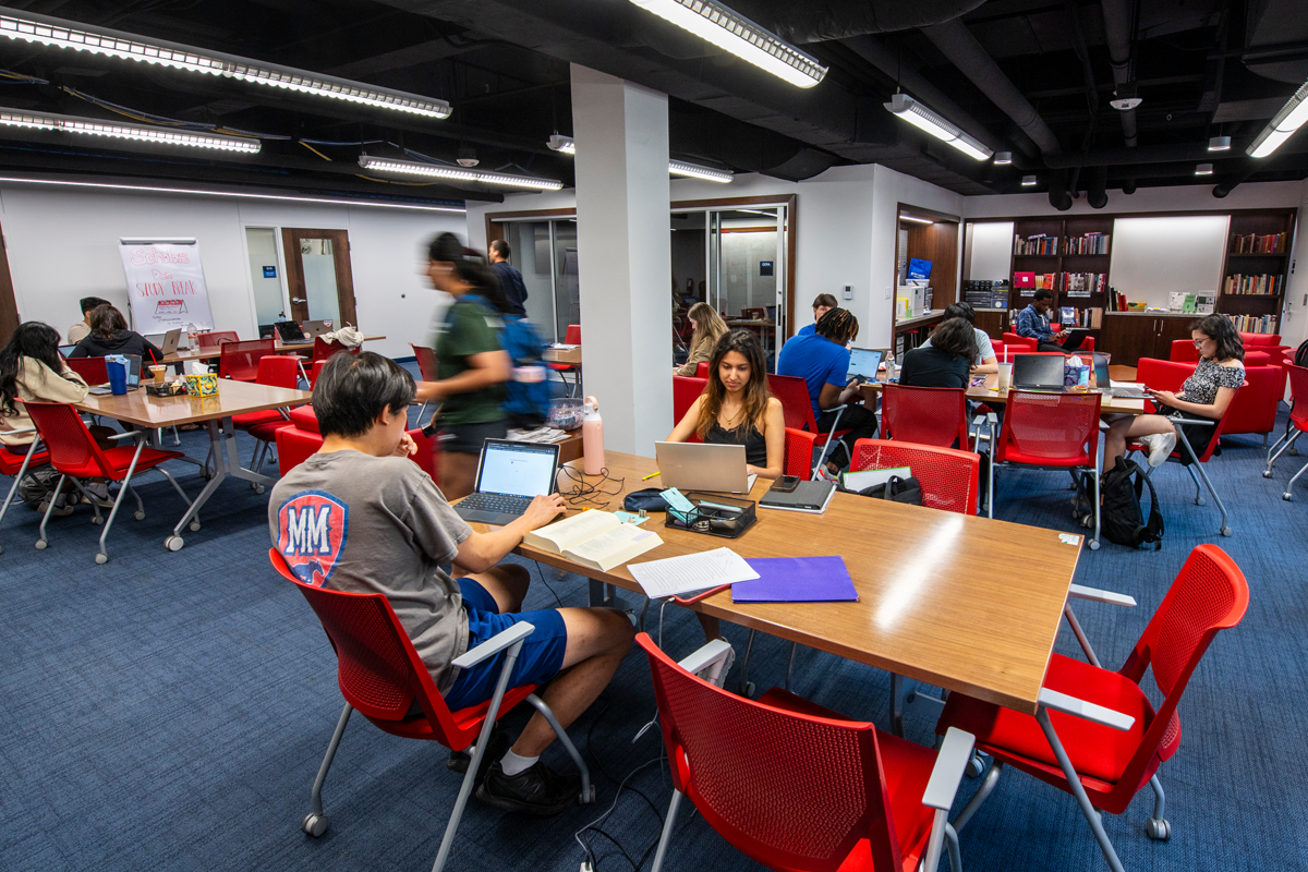 Students studying and working on laptops in a bright, modern Scholars Den space with red chairs and shared tables.