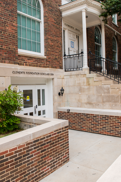 Exterior entrance to the Clements Foundation Scholars Den in the lower level of Clements Hall, featuring red brick walls, white trim, large arched windows, and a ramp leading down to glass double doors.