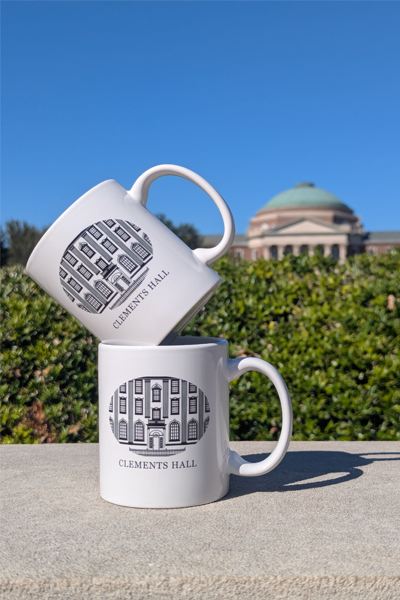 Two white “Clements Hall” mugs, one stacked at an angle on top of the other, sit on a ledge outdoors with green hedges and a domed campus building under a clear blue sky and the top of Dallas Hall in the background.