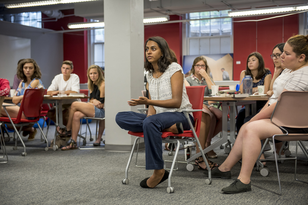 Students are seated at tables in the Scholars' Den, while one student asks a question to a professor who is out of the camera's frame.