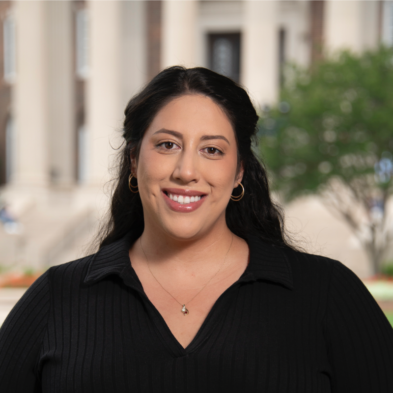 A woman with long, wavy dark hair and a warm smile. She is wearing a black, ribbed sweater with a collared neckline, gold hoop earrings, and a delicate gold necklace. The background is softly blurred, showing steps, greenery, and classical-style columns of a university building.
