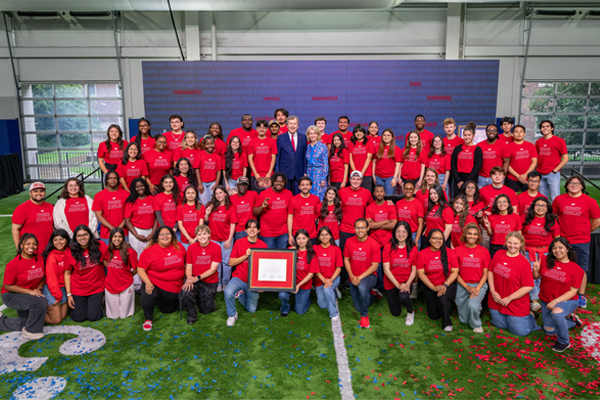 A large group of students and staff wearing matching red “Turner Scholars” T‑shirts pose together on an indoor turf field, standing and kneeling in several rows around a framed certificate, with Dr. Gerald and Gail Turner dressed formally in the center and a blue word‑wall backdrop behind them.