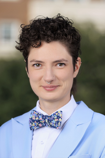 Alexandra Thibeaux is a white woman with short curly hair, cut in a pixie style. She is wearing a light blue suit jacket, white dress shirt, and a colorful floral-patterned bow tie. The background is the blurred path towards Dallas Hall.