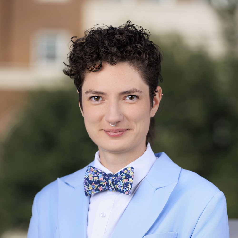 Alexandra Thibeaux is a white woman with short curly hair, cut in a pixie style. She is wearing a light blue suit jacket, white dress shirt, and a colorful floral-patterned bow tie. The background is the blurred path towards Dallas Hall.
