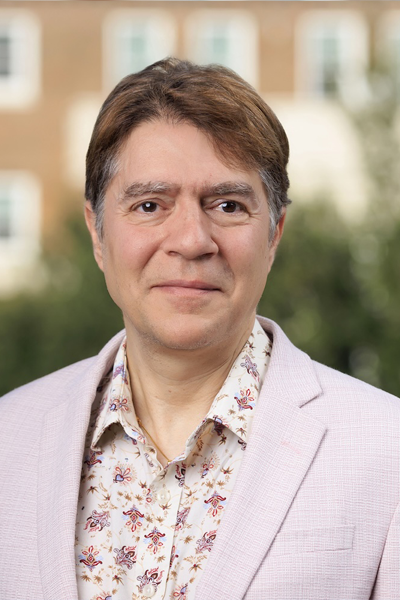 Dr. Barr is a light complected man with short brown hair is smiling at the camera. He is wearing a light-colored blazer over a patterned button-down shirt. He is photographed outdoors in front of a softly blurred building and greenery.