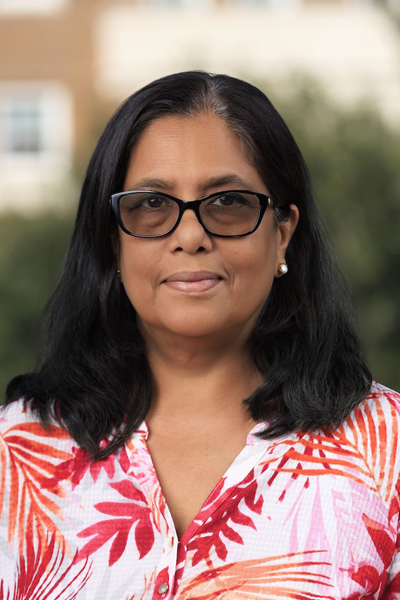 Dr. Madhavi Biswas has straight, shoulder-length black hair and medium tan skin with warm undertones. She is wearing dark-framed glasses and a button-down shirt with a red and pink tropical leaf pattern, looking calmly at the camera. Th background is outdoors in front of a softly blurred background of trees and a building
