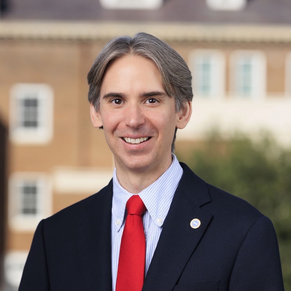 Dr. Brandon Gray Miller is a white man with straight, medium-length silver and brown hair. He is wearing a dark suit jacket, light blue striped dress shirt, and bright red tie, posed smiling in front of a softly blurred outdoor background with a building and greenery.