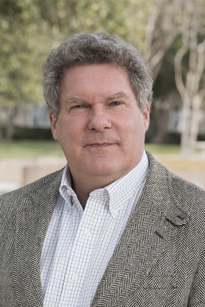 A man with short curly gray hair, wearing a gray herringbone blazer over a white and blue checkered dress shirt. He is standing outdoors in a blurred background of trees and a college campus, facing the camera with a neutral expression.