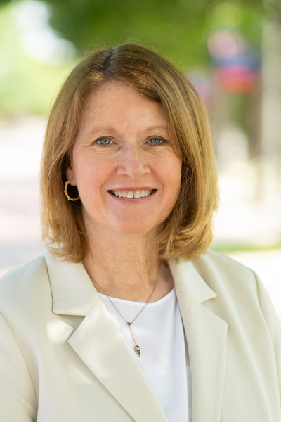 Susan is a white woman with shoulder-length dark blonde hair. She is wearing a beige blazer with a white blouse underneath. The background is the blurred path to Dallas Hall.