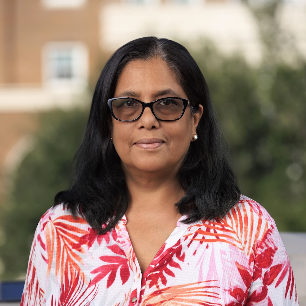 Dr. Madhavi Biswas has straight, shoulder-length black hair and medium tan skin with warm undertones. She is wearing dark-framed glasses and a button-down shirt with a red and pink tropical leaf pattern, looking calmly at the camera. The background is outdoors in front of a softly blurred background of trees and a building