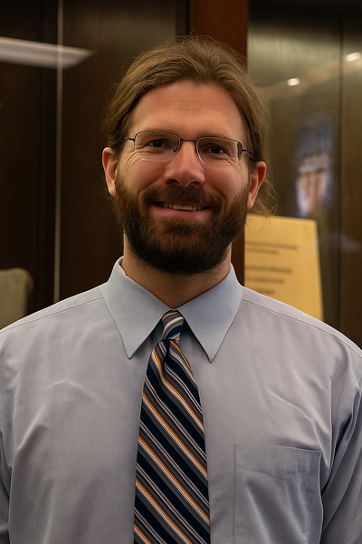 Dr. Russ McConnell is a white man with long brown hair pulled back, a full beard, and glasses smiles while standing in front of a dark wooden display case. He wears a light blue dress shirt with a multi-colored striped tie. 