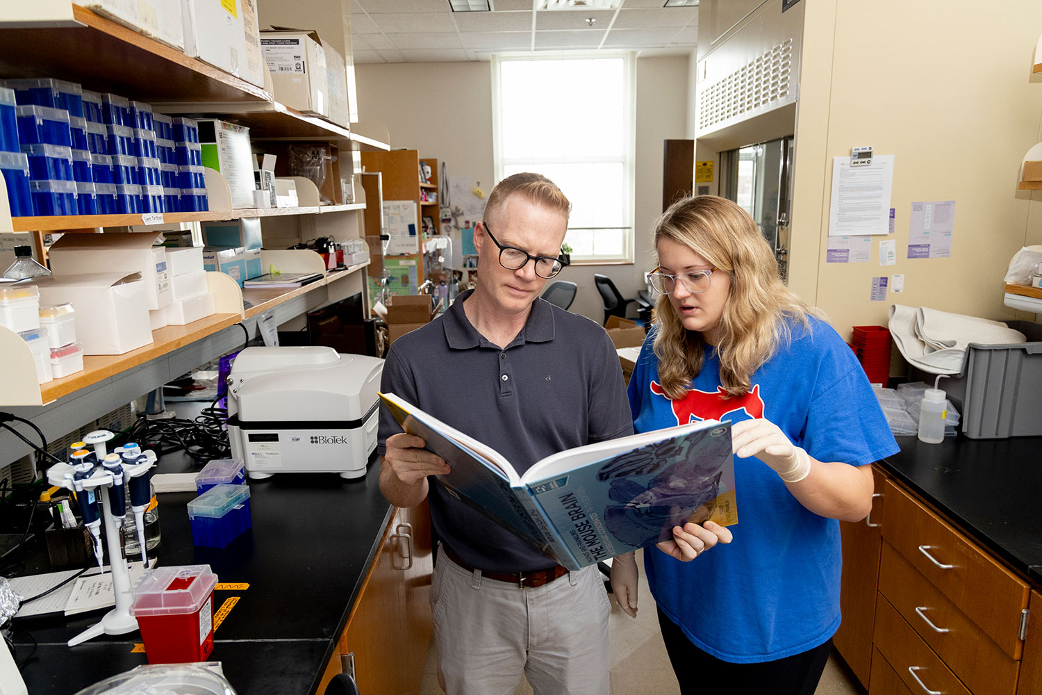 Two people in a laboratory reviewing a large book together beside lab equipment and pipettes.