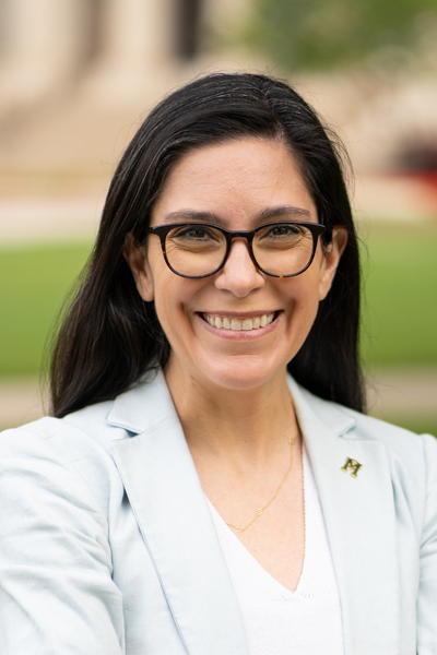 Dr. Caitlin Anderson is a white woman with straight dark brown hair past her shoulders. She wears a white v-neck blouse under a white blazer. She also wears black framed glasses.
