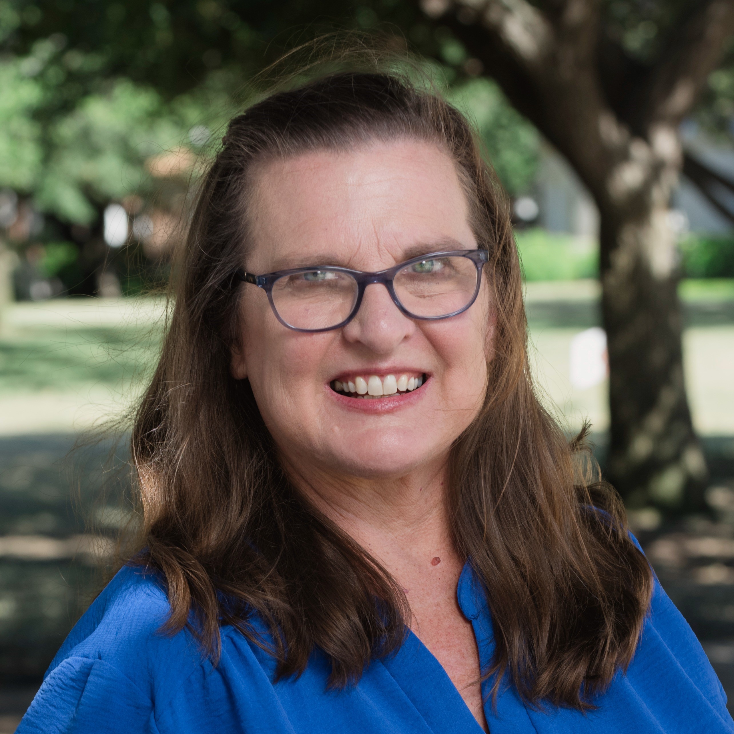 A woman with long brown hair and black framed glasses smiles warmly while standing outdoors. She is wearing a bright blue blouse, and the background features green trees and a sunlit lawn.