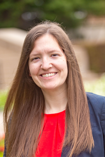 A Caucasian woman with long straight brown hair smiles at the camera. She is wearing a bright red top and a navy blazer. The photo is taken outdoors with a soft-focus background of greenery, flowers, and a stone path. The lighting is natural and warm.
