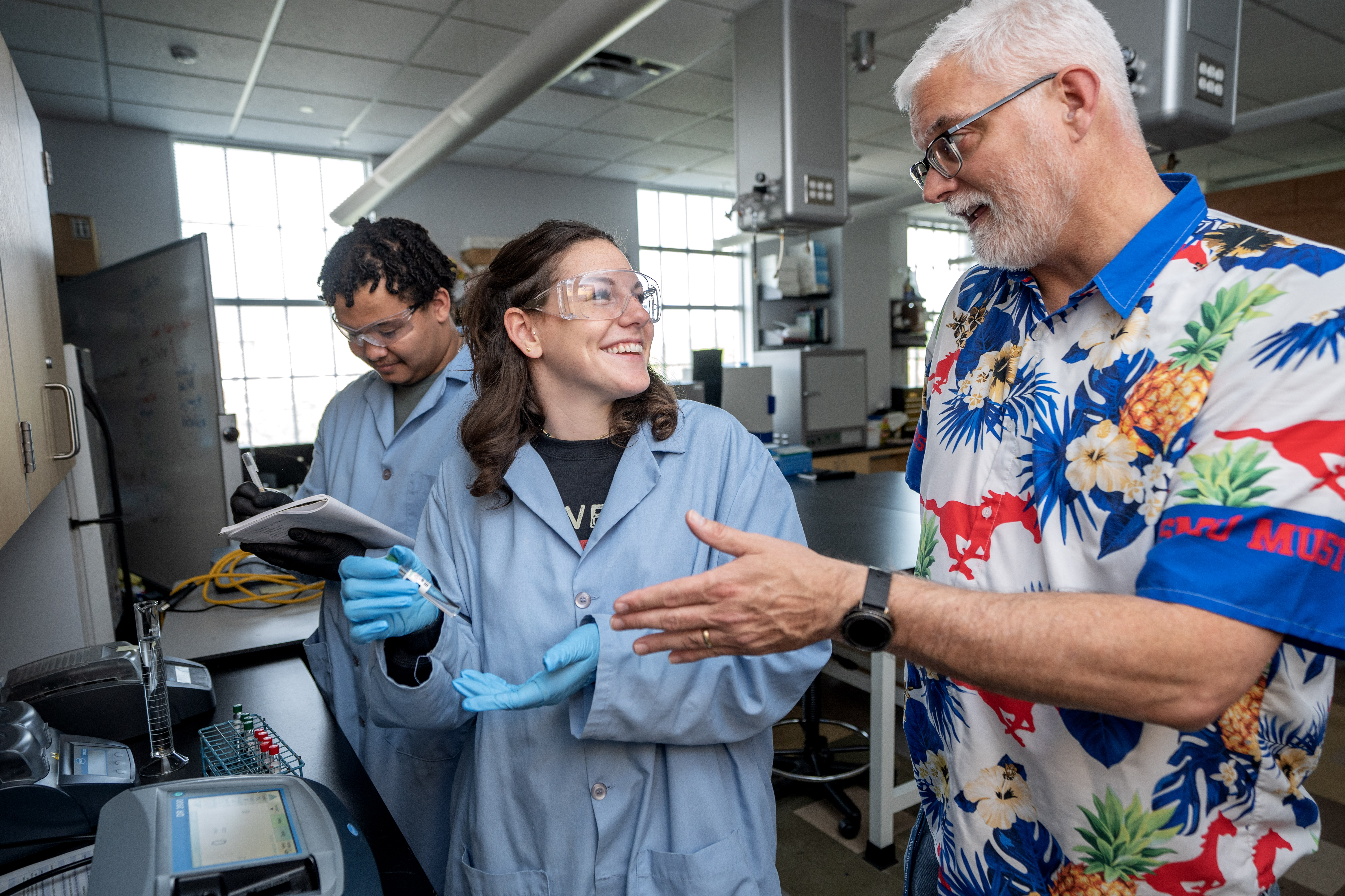 Students wearing lab coats in a lab receiving instruction from a professor
