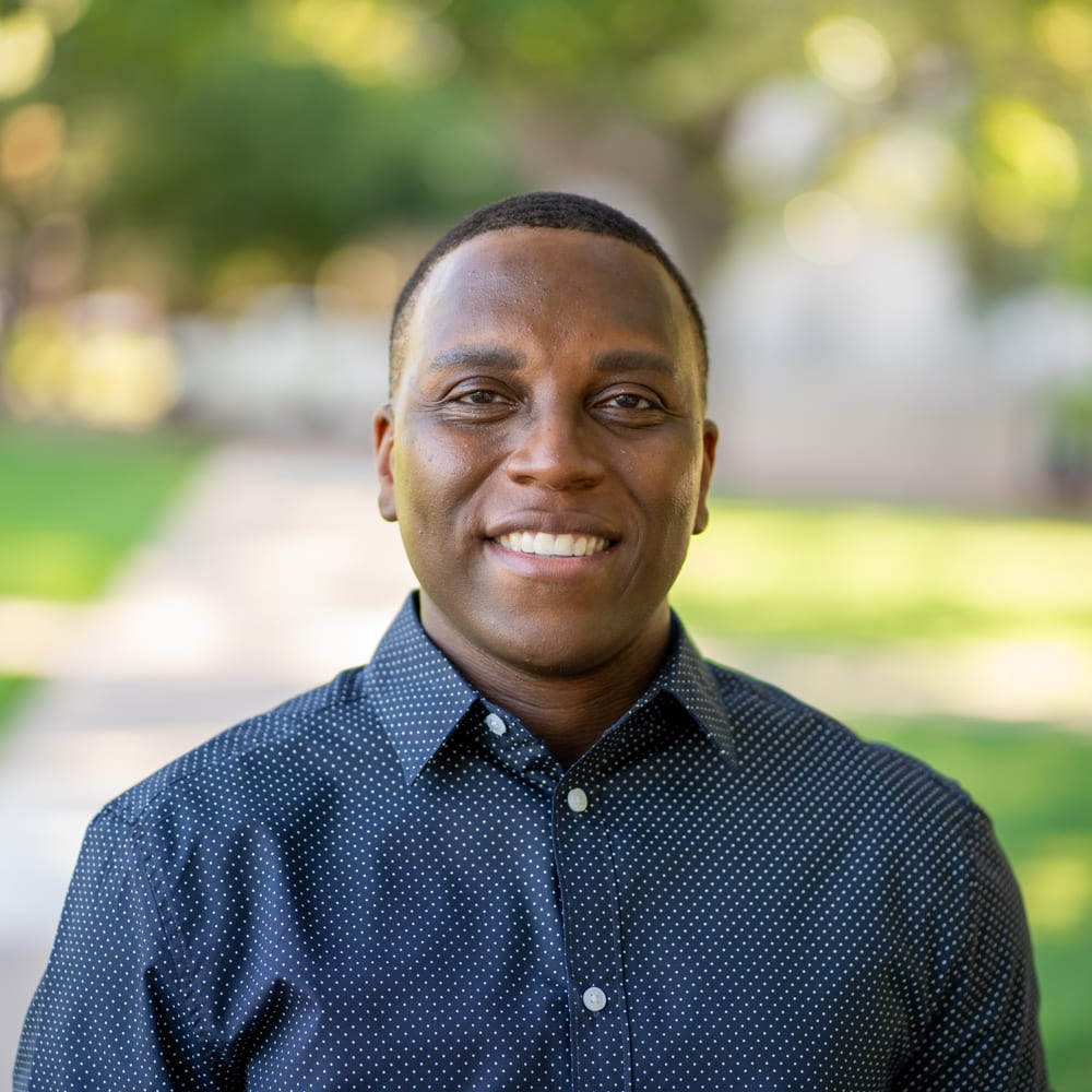 Photo of Shun Colter smiling, standing on SMU campus with a path and trees behind him