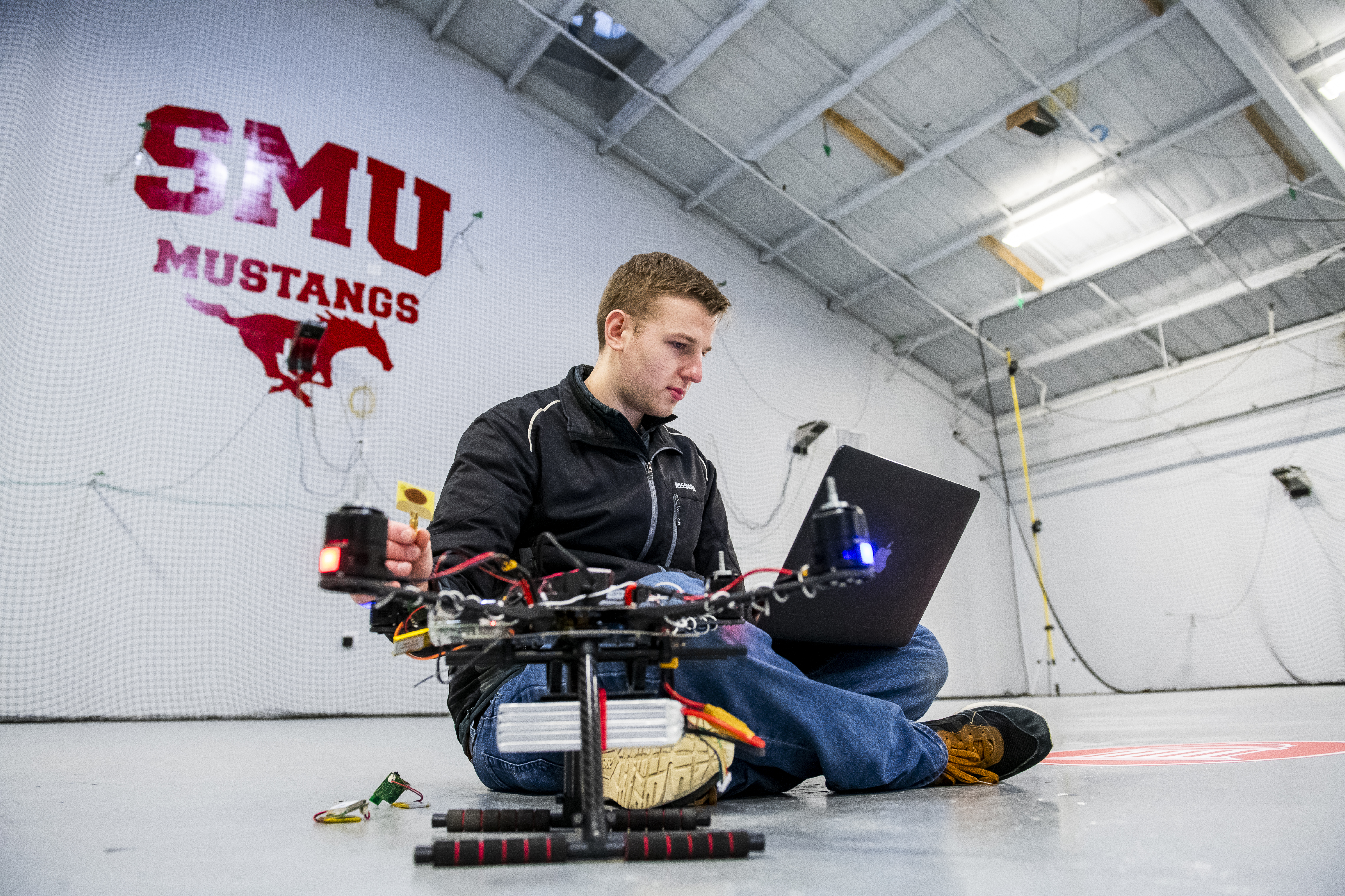 student sitting on floor with laptop working on remote control drone next to him