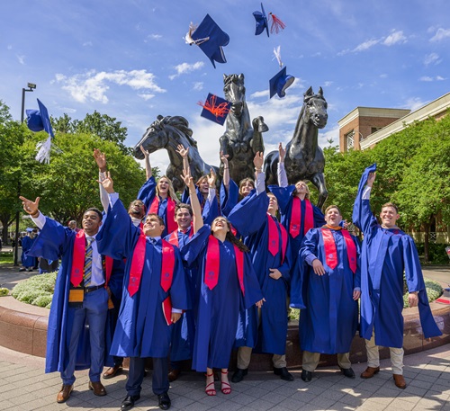 graduating students throwing mortar boards into the air in front of a bronze mustangs statue