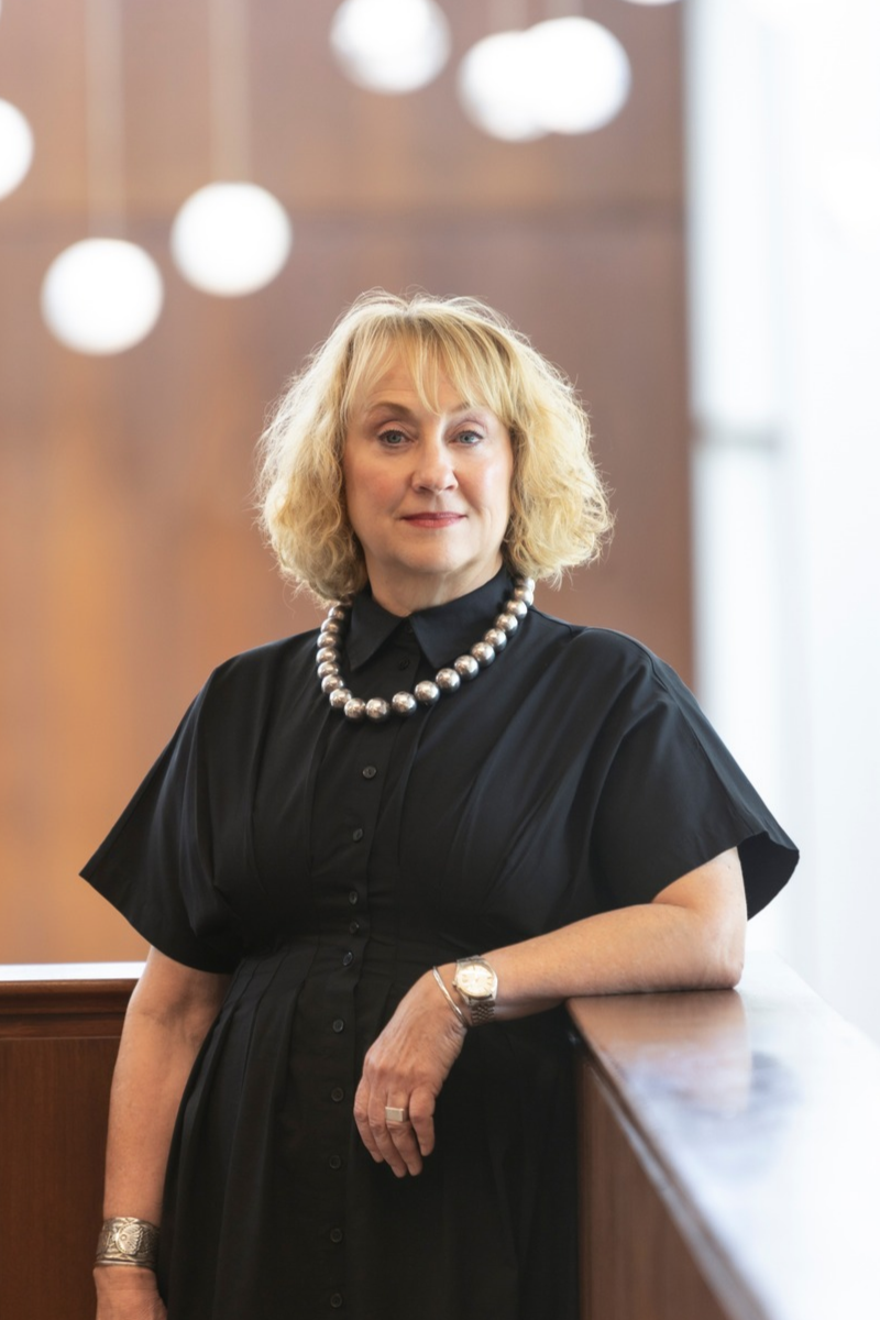 Melissa Chessher has short blonde hair, and is wearing a black dress with a large pearl necklace. She is posed in the Owen Arts Center Balcony looking into the Jones Atrium.