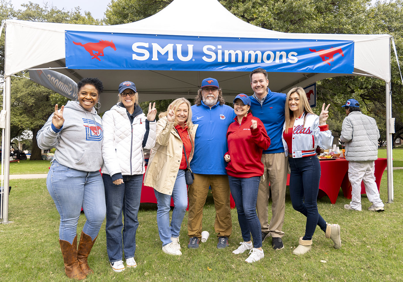 Attendees pose beneath an SMU Simmons-branded tent during the Simmons Homecoming Tailgate, gathered on a lawn near tailgate tables and activities before an SMU game.