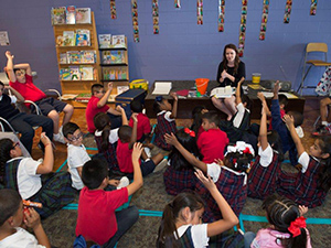 A group of children sitting on the floor, raising hands, engaged with a teacher in a classroom setting.