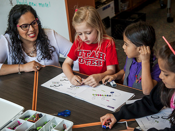 A teacher guides students as they collaborate on a project at a classroom table filled with books and materials.