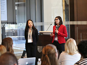 Dr. Denisa Gandara stands with a colleague before a large audience at a conference.