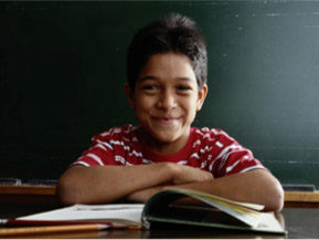 A smiling boy sitting at a desk, focused on reading a book.