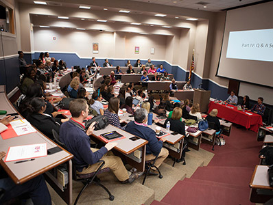 Interior of a spacious lecture hall with numerous attendees sitting in chairs, engaged in a presentation.