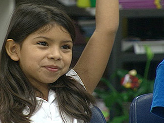 A young girl raises her hand in class, eager to participate and answer a question posed by the teacher.