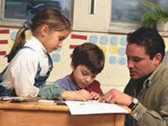 Two children sit at a desk with a teacher, engaged in a reading intervention activity.