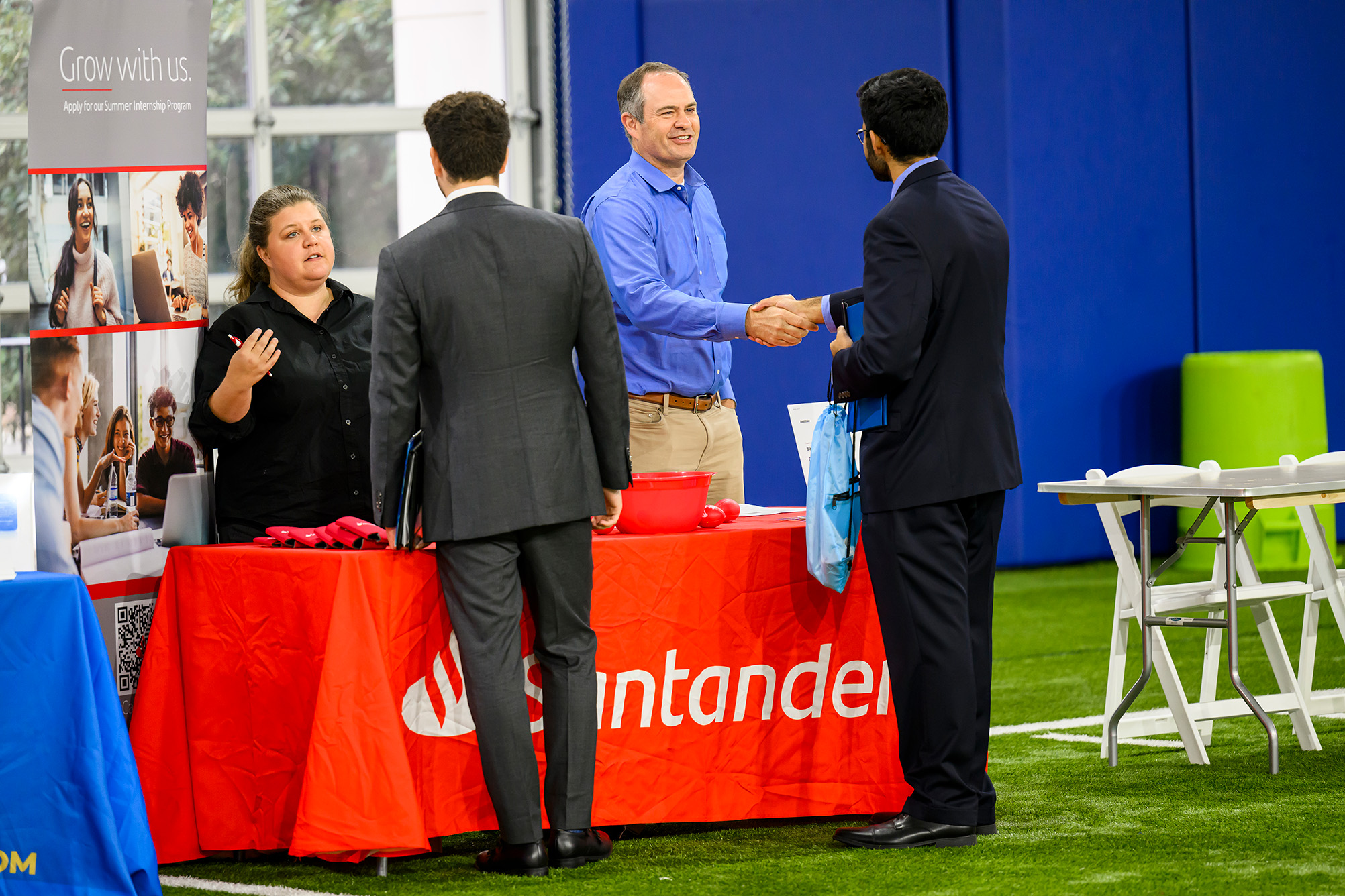 two people standing at table facing two people on other side of table talking, table has red cloth with white business logo, window in background and standing on a green turf field