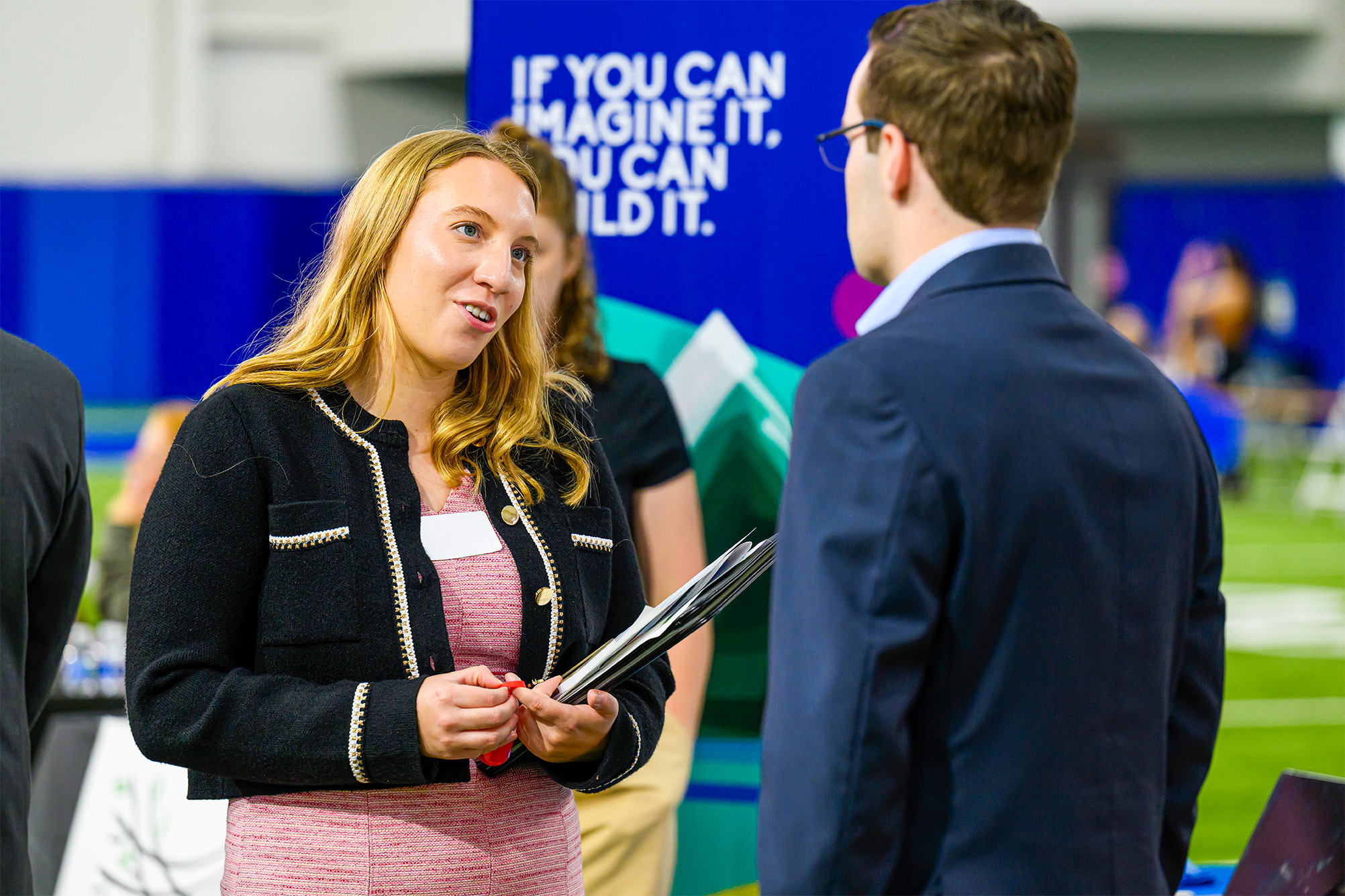 woman in pink dress with black blazer jacket standing and talking to man in blue blazer jacket