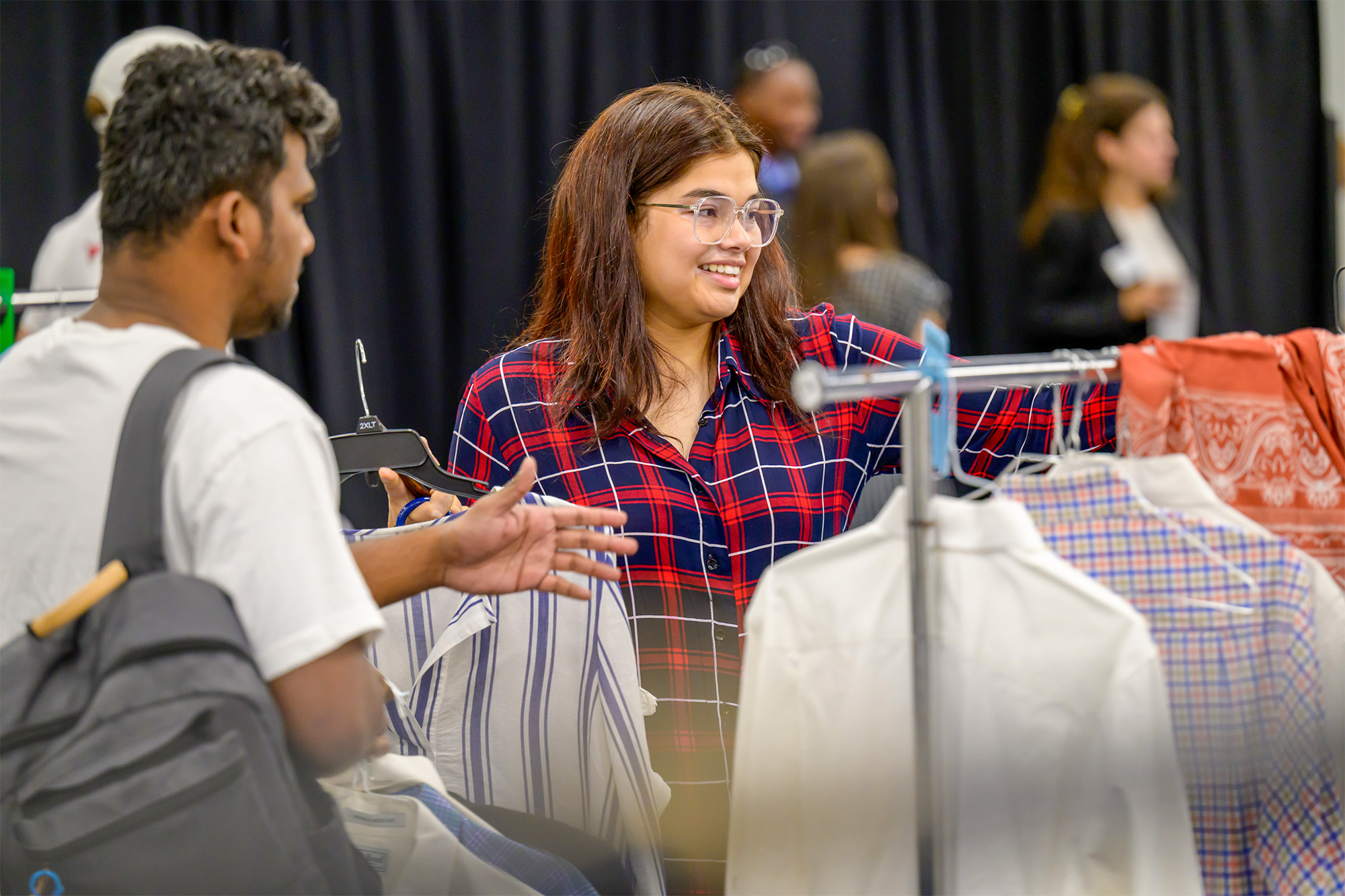 student in blue and red plaid shirt  with student in white shirt and gray backpack standing by clothes hanging on a rack