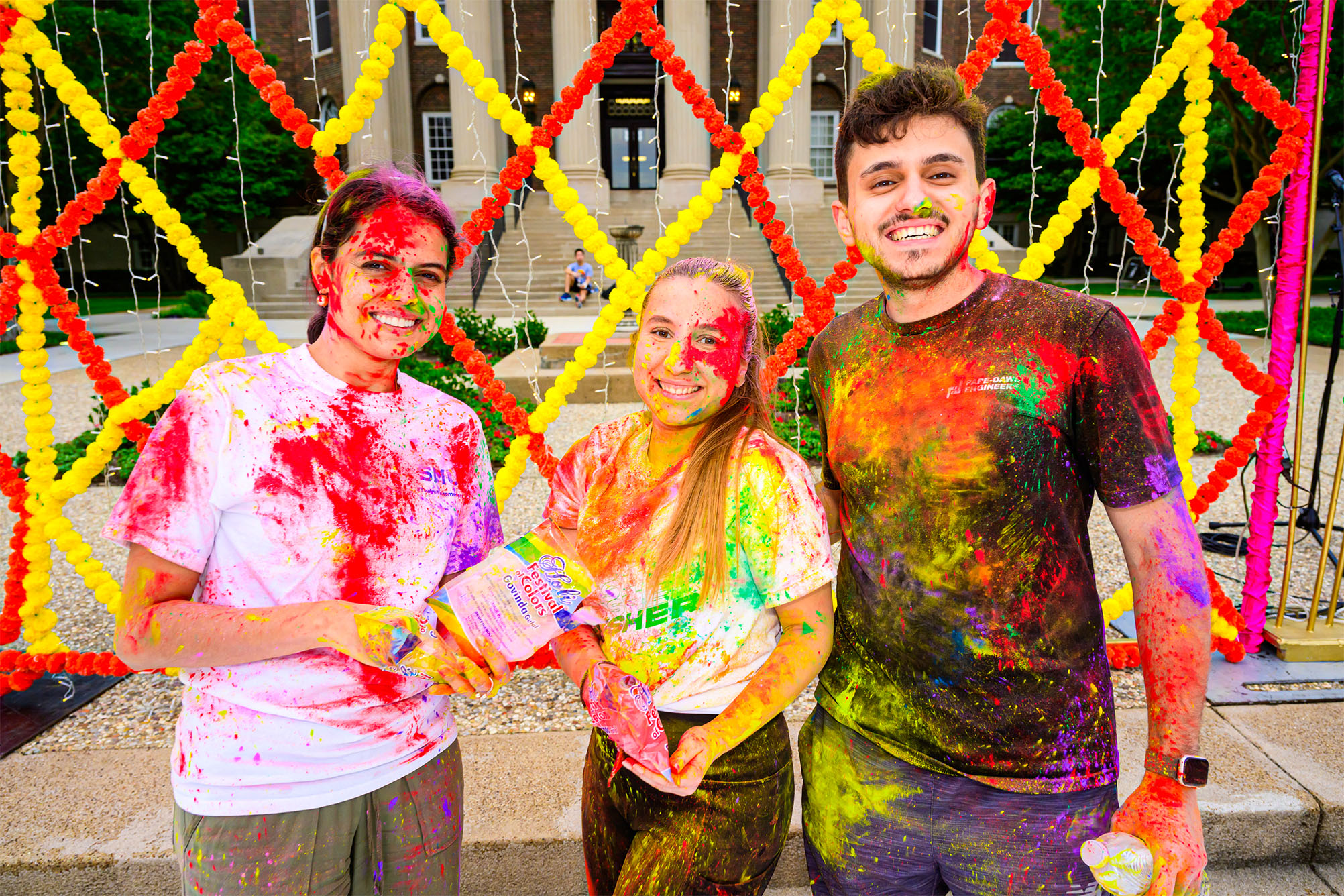 three people covered in multi-colored powder standing outside in front of red and yellow floral backdrop