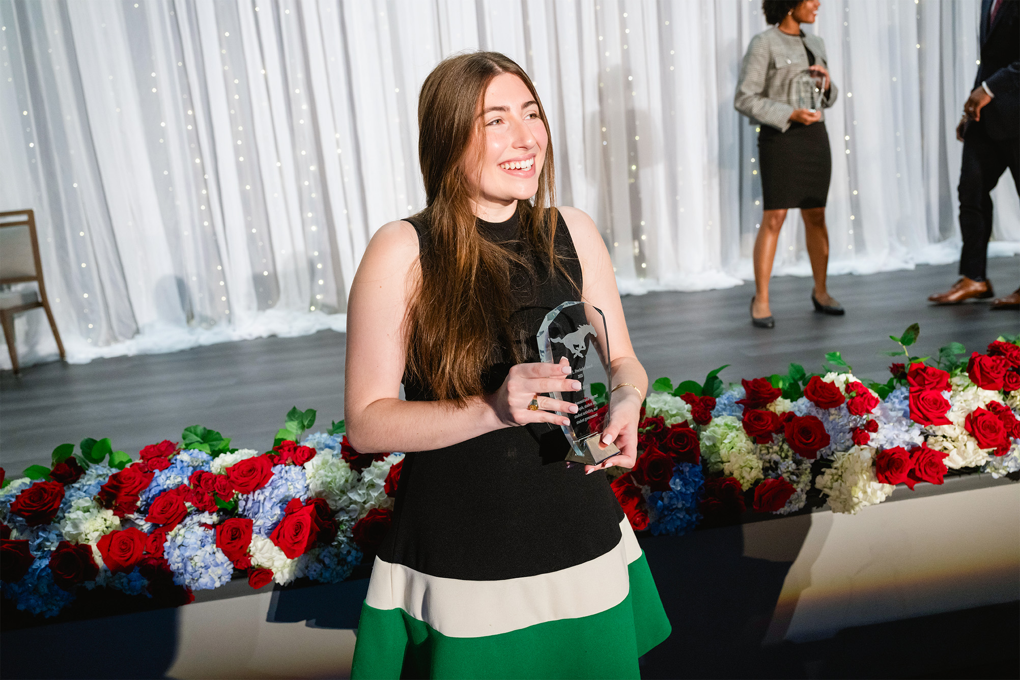 student posing for photo with crystal award plaque in front of stage with red, blue, and white flowers in the background