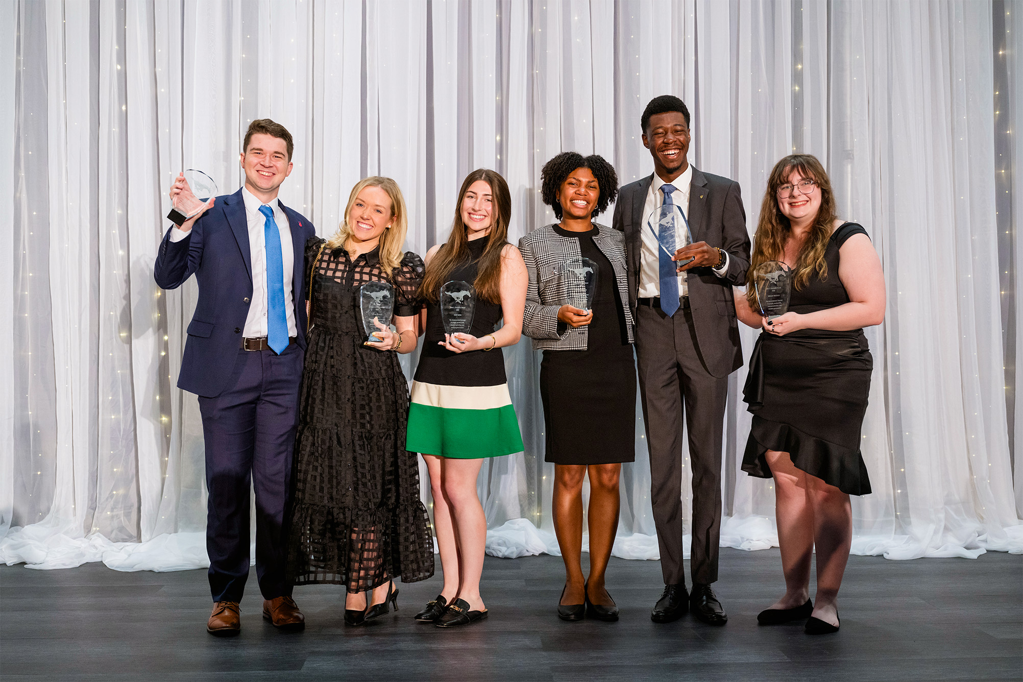 six students standing on stage with crystal awards posing for photo with white curtain backdrop