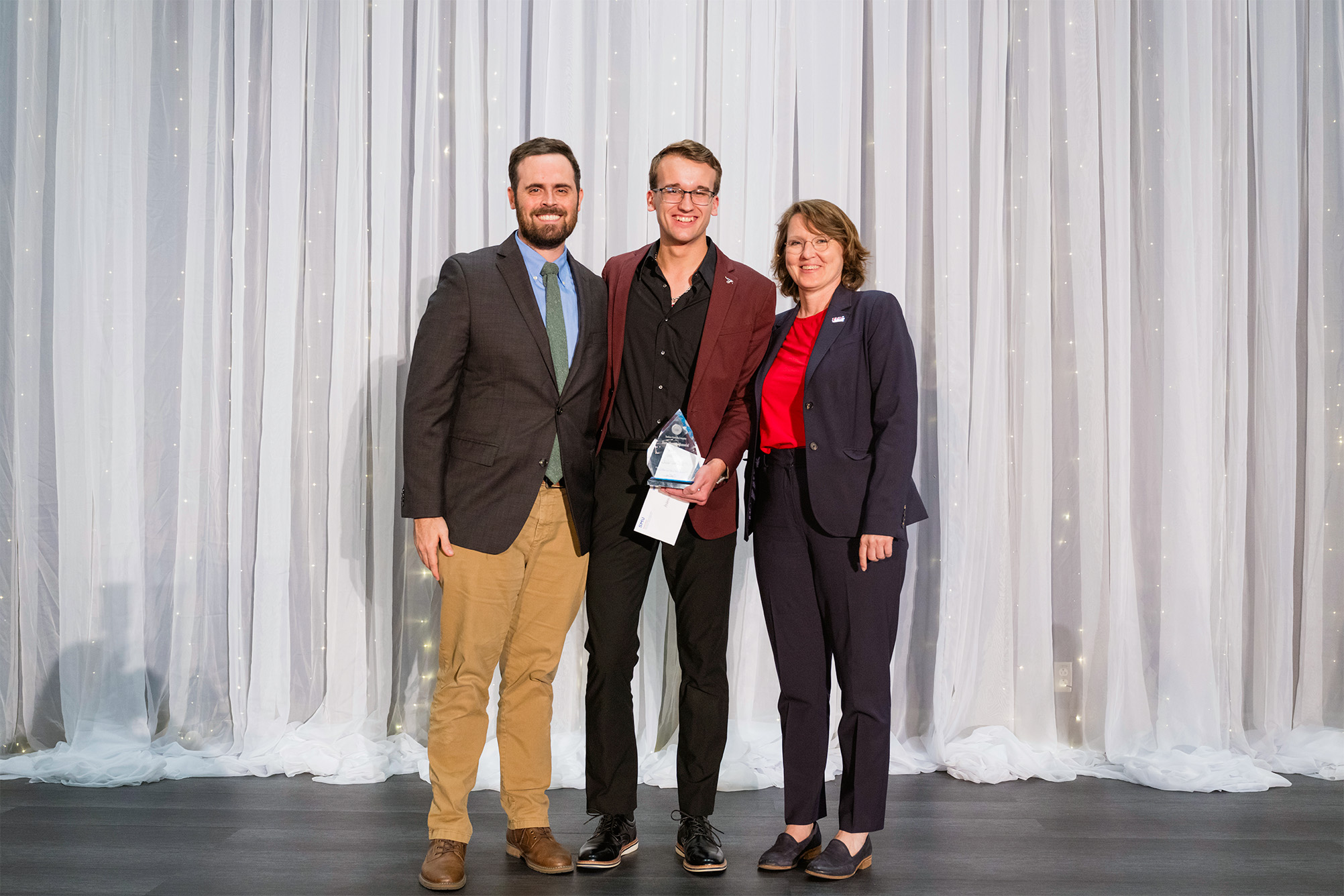 three people posing for photo on stage with award recipient in the middle holding award plaque