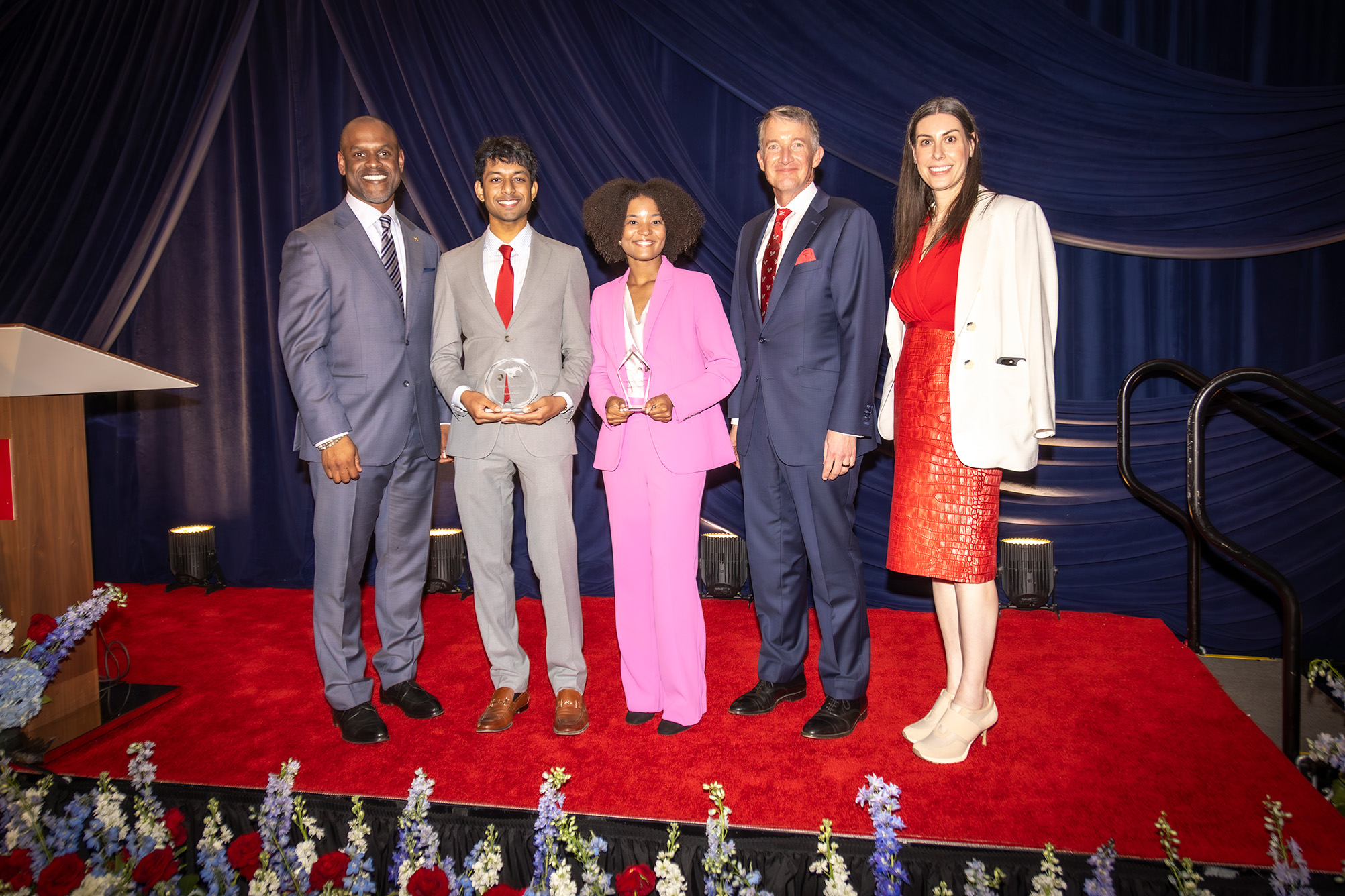 five people standing on stage posing for photo with dark blue drapes behind and red carpet on the stage
