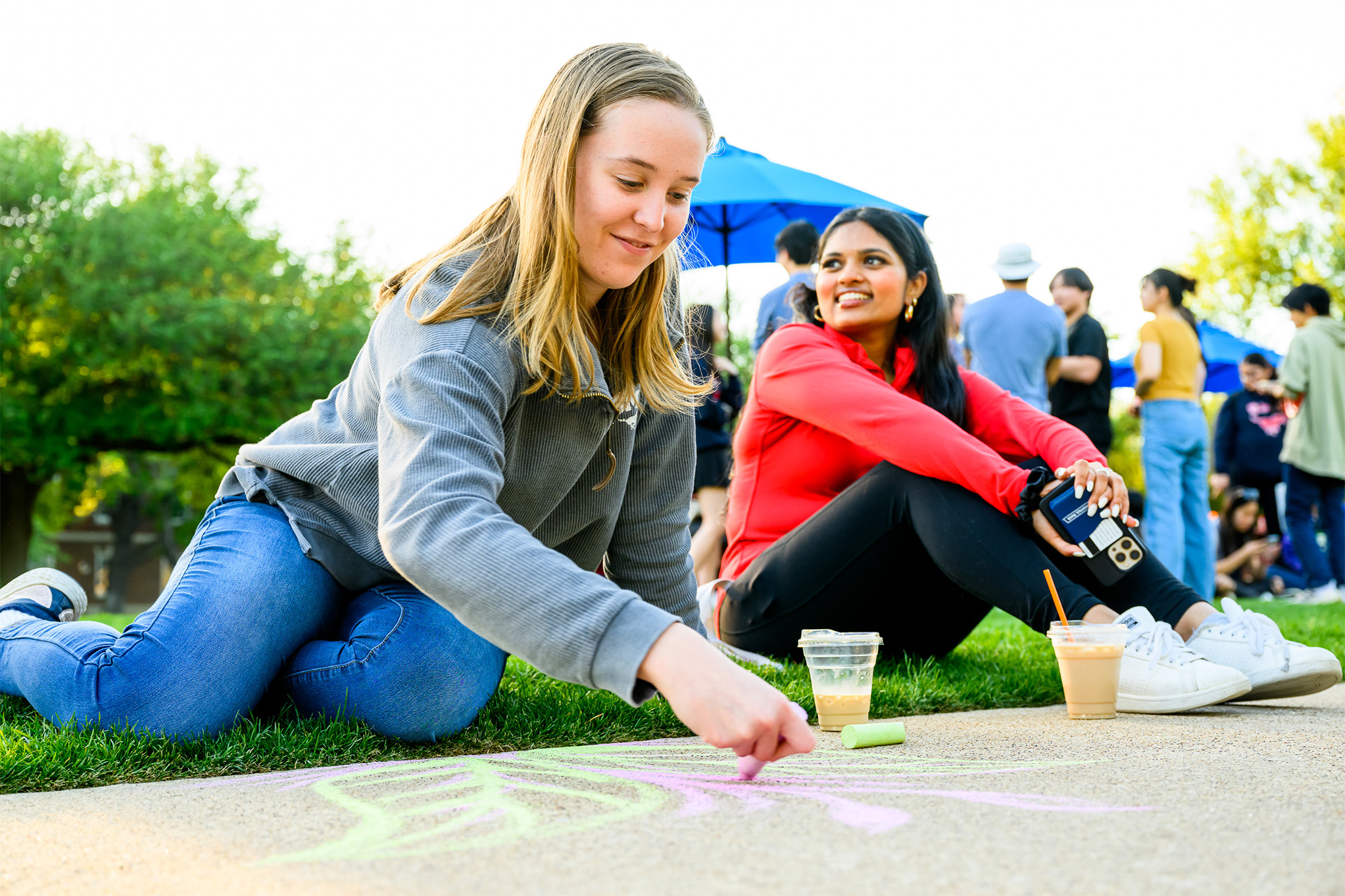 two students sitting on sidewalk, one drawing with chalk