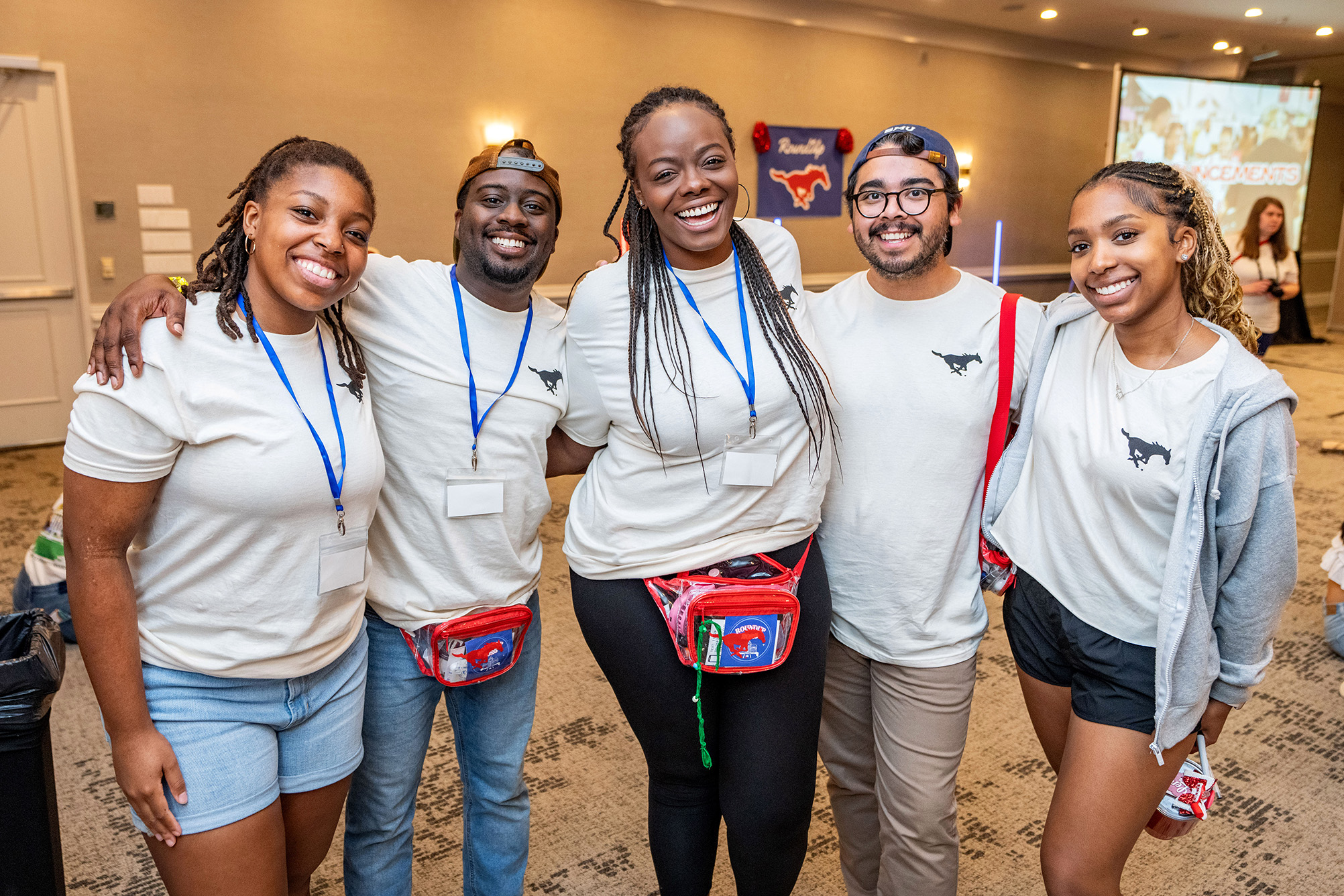 five people standing together indoors for a photo, smiling, and wearing white tshirts with a black mustang logo